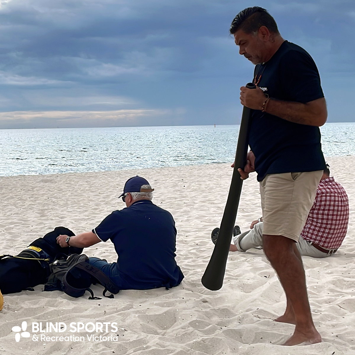 Lionel, now with shoes off like the BSRV participants, is standing holding the Yikadi (digeridoo) in his hands looking downwards as though in contemplation. There are people sitting on the beach including one person with their Guide Dog.